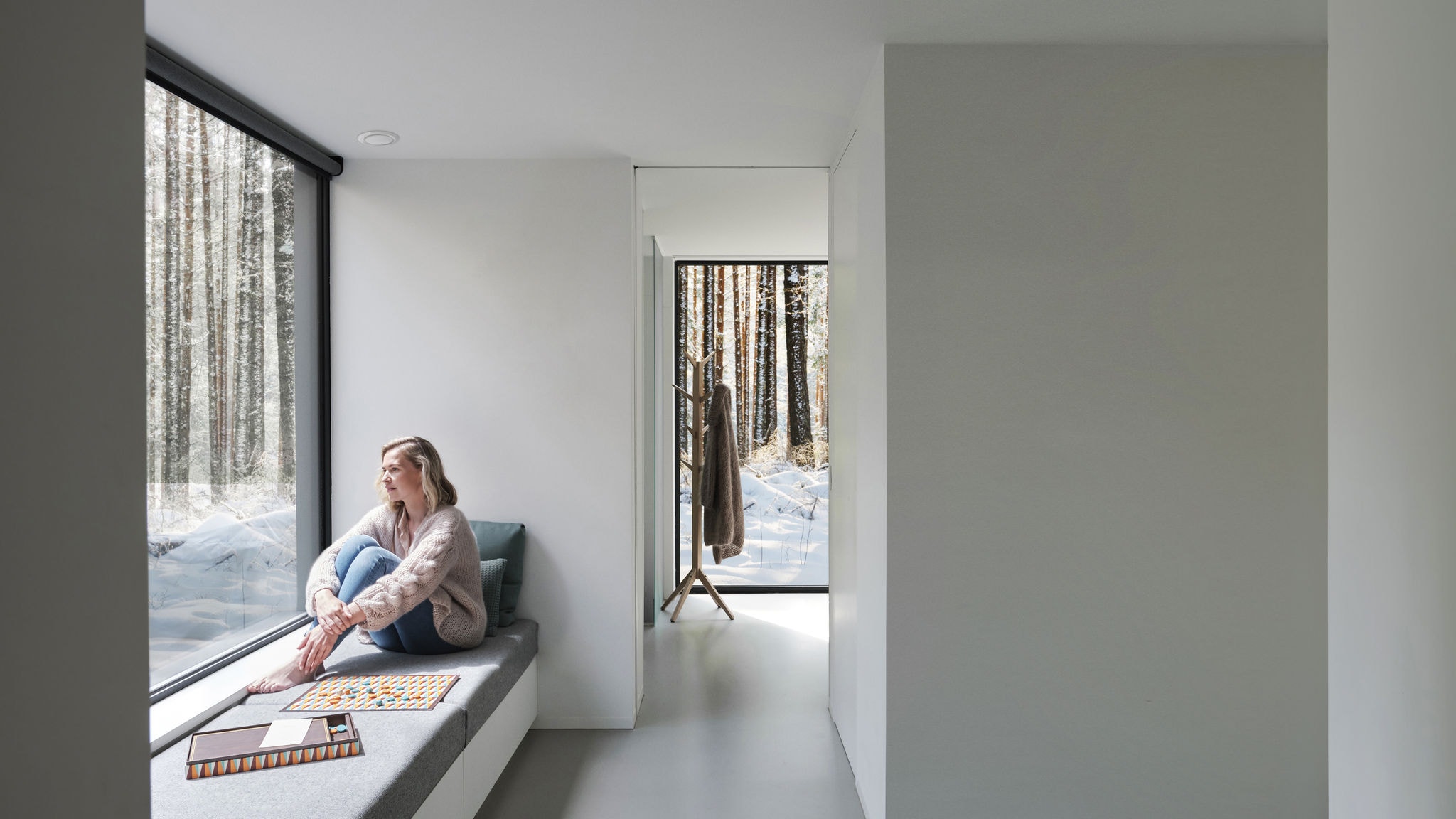 Wide shot of woman sitting in front of a window barefoot, looking out towards the trees and the snow.