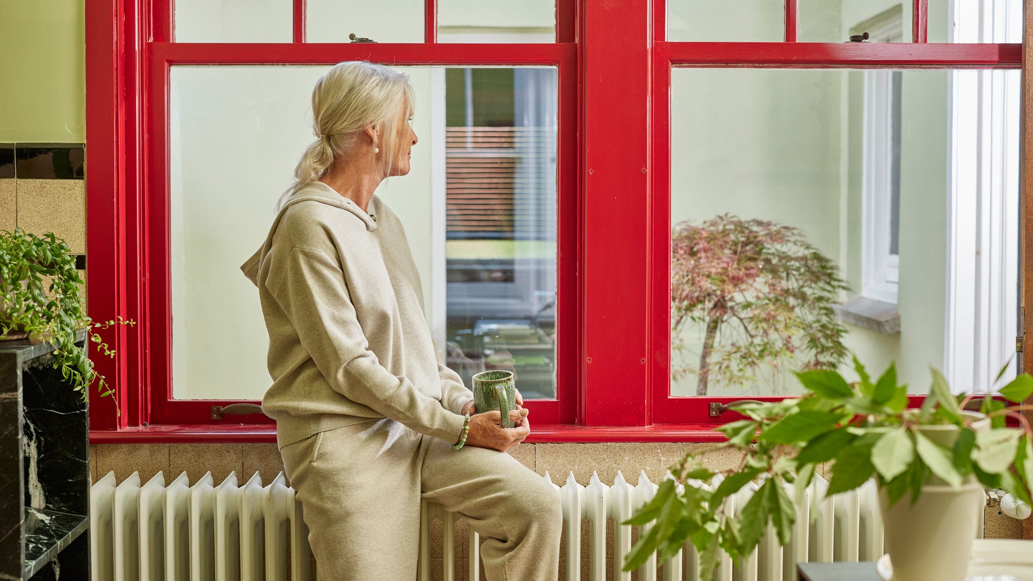 Woman sitting on a radiator by a large window, enjoying a warm, energy-efficient home