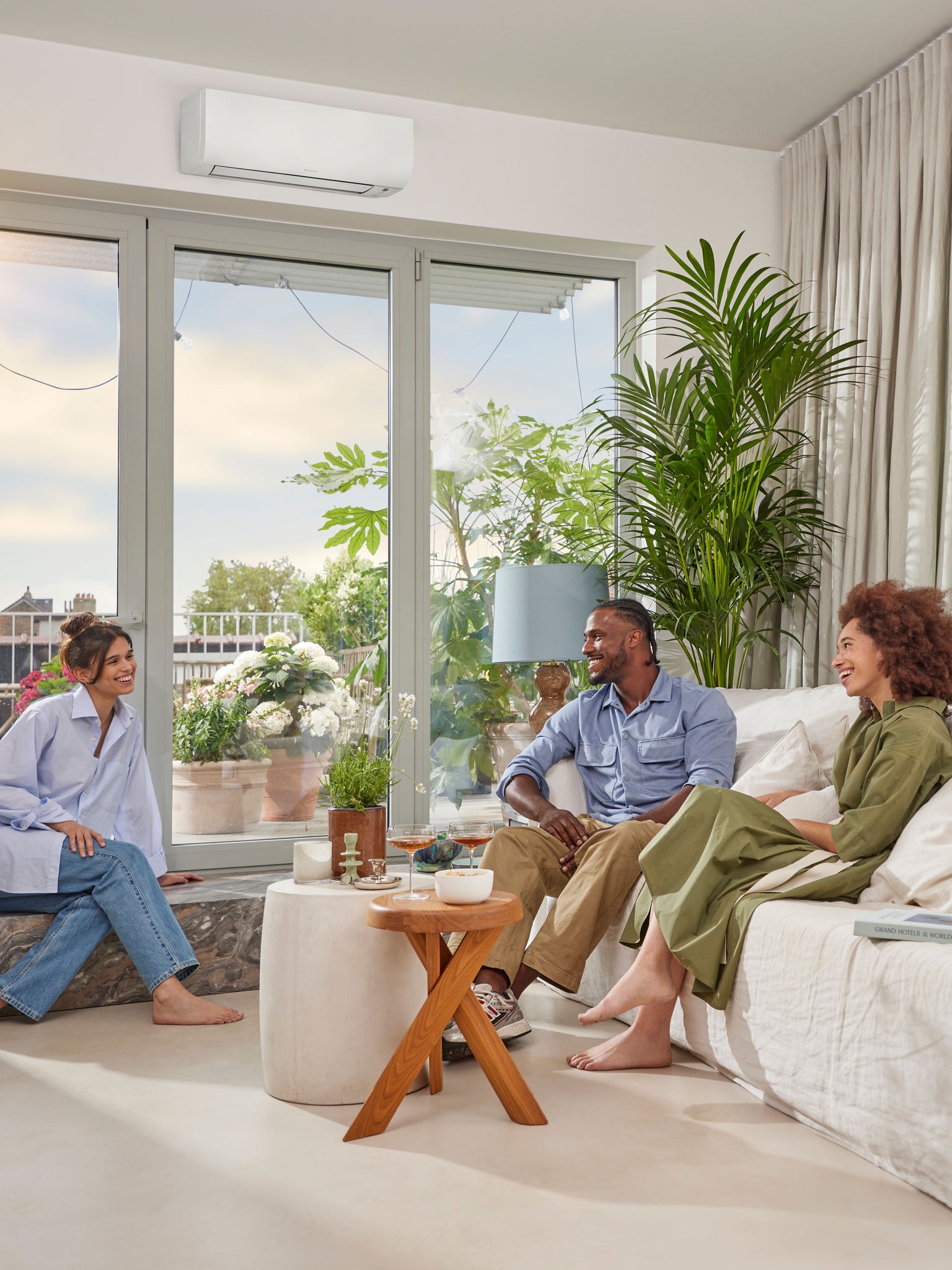 A man and two women sitting and chatting in a bright, airy room with white walls and a discreet, white, wall-mounted heat pump above a large picture window on a sunny day