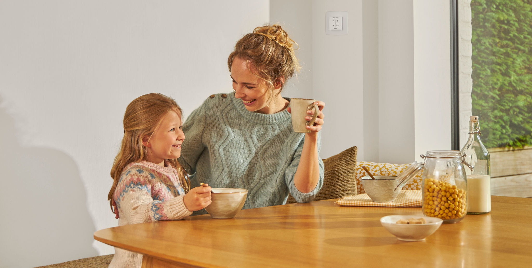 Young girl eating from a bowl seated at a table next to a woman with a mug in hand, both smiling, happy and relaxed, enjoying healthy air.