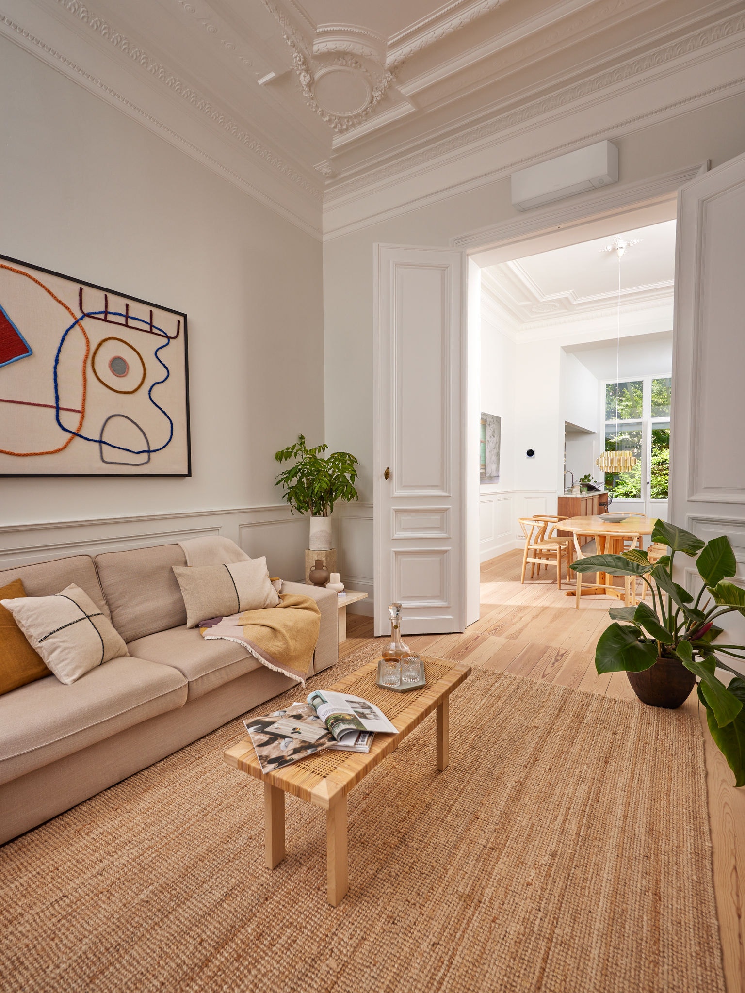 White, wall-mounted heat pump above a doorway leading from comfortable sitting room with a cream sofa into a dining room bathed in light from a large window