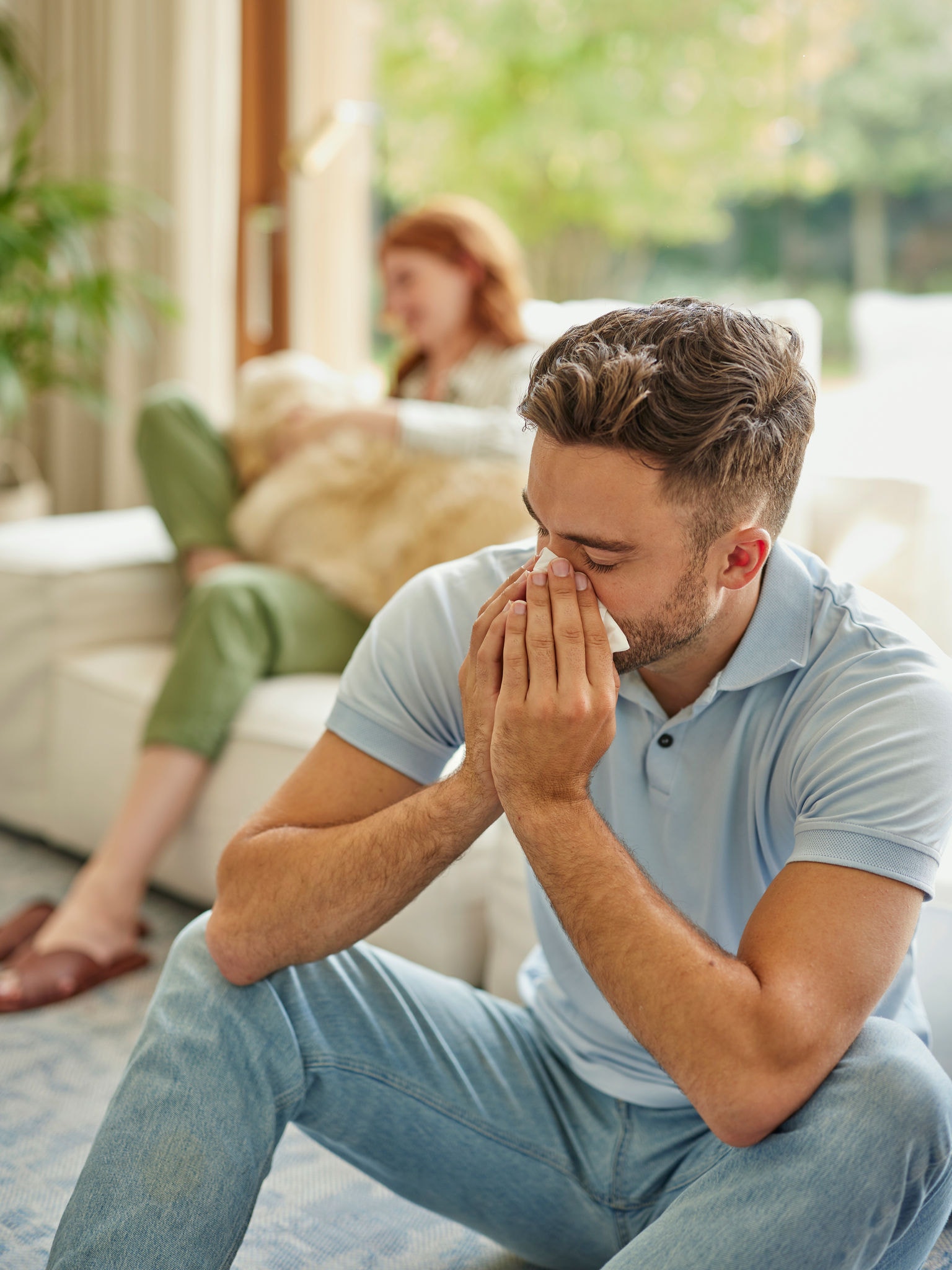 Woman in background on soft furnishings with golden retriever on lap, man in foreground sneezing